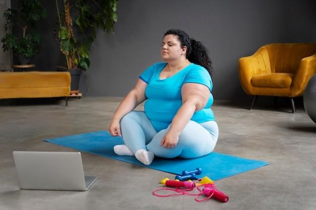 Woman in workout clothes sitting on a yoga mat with dumbbells and jump rope, exercising at home.