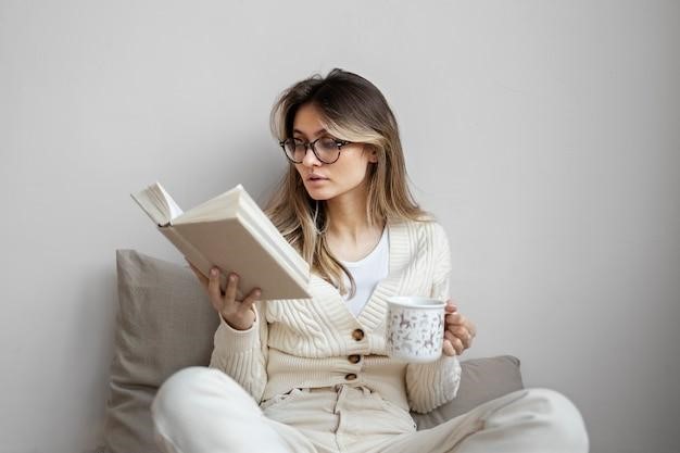 Woman sitting comfortably while reading a book and holding a mug, staying informed and relaxed.