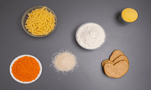 Flat lay of carbohydrate foods including dry pasta, flour, rice, red lentils, bread, and cornmeal.