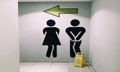 Restroom signage with male and female pictograms crossing their legs under a directional arrow.