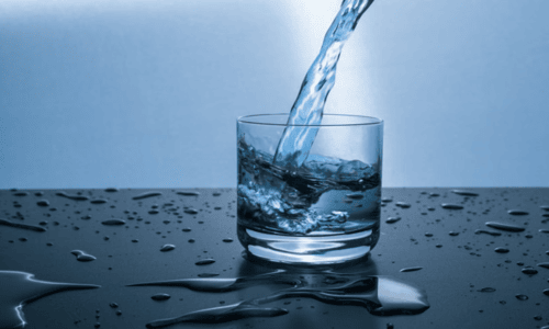 Water pouring stream filling a clear glass surrounded by fresh water droplets on a dark surface.   