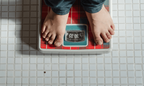Bare feet standing on a red plaid analog bathroom scale placed on white tiled floor.
